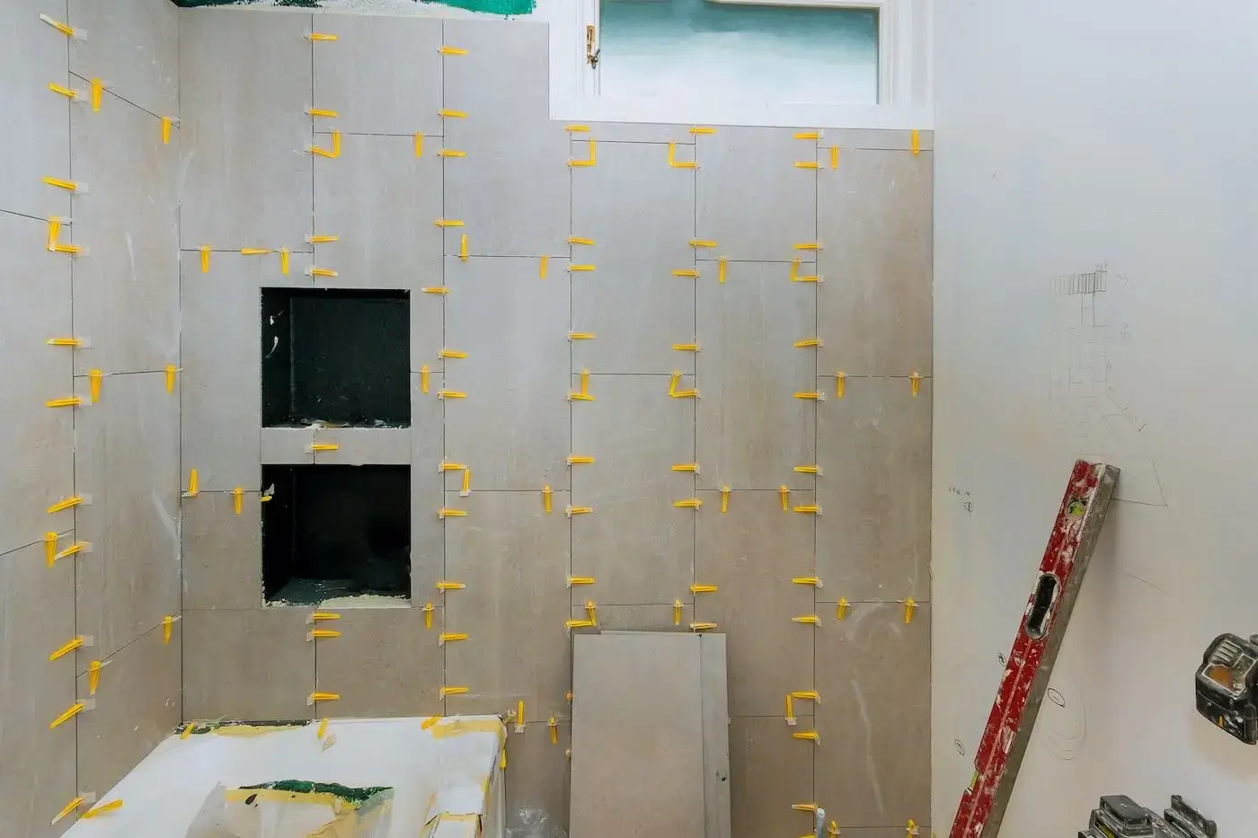 Large-format wall tile going up around a recessed shower niche during a bathroom remodel, with yellow levelling clips still in place